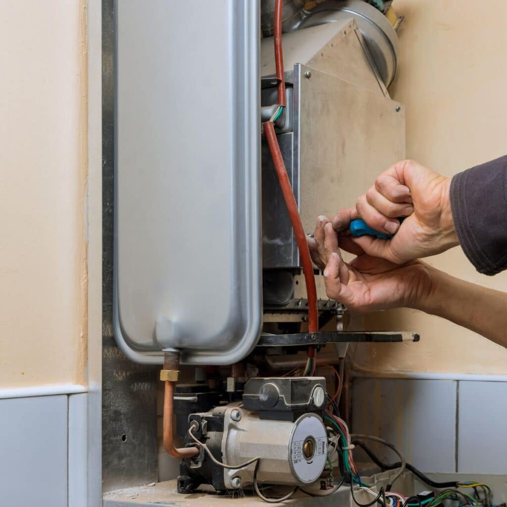 A person is servicing a boiler or water heater, visible by their hands using a screwdriver to adjust components within the lower service panel. The complexity of the machinery, with various wires and pipes, suggests technical expertise is required for the task. The maintenance work is being performed in a domestic setting, indicated by the tiled wall and flooring