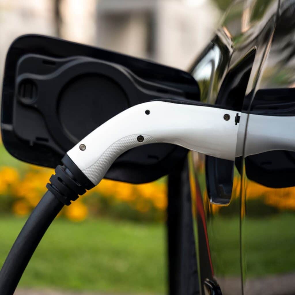 A close-up image of a white and black electric vehicle (EV) charging cable connected to the charging port of a car, with the car's reflective surface mirroring a serene outdoor setting. The charger is securely fastened, indicating that the vehicle is actively charging. The open charge port cover and the cable's positioning suggest a focus on modern EV technology and sustainable transportation
