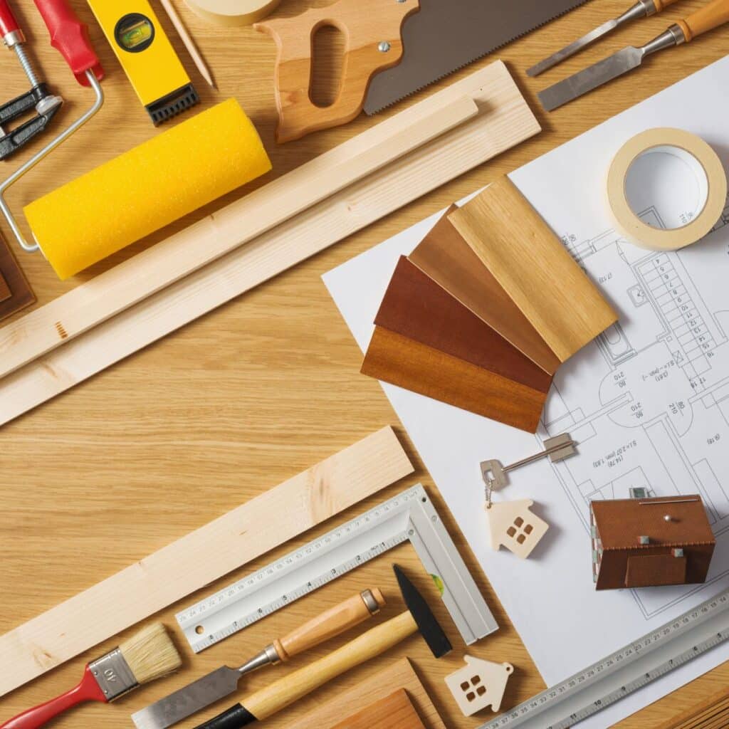 A variety of carpentry and home improvement tools spread out on a wooden surface alongside architectural blueprints. Items include a handsaw, paint roller, measuring tape, wood planks, chisel, and brush, indicating an ongoing construction or DIY project. The arrangement of tools and samples suggests meticulous planning and manual work