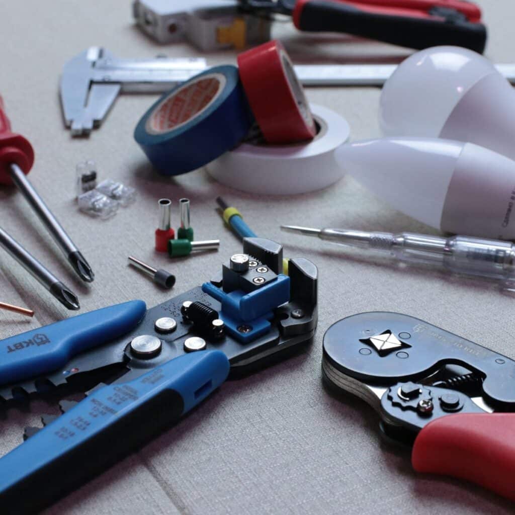 An array of electrical tools and components laid out on a fabric surface, including wire strippers, screwdrivers, cable cutters, and various connectors, indicative of an electrical repair or installation project