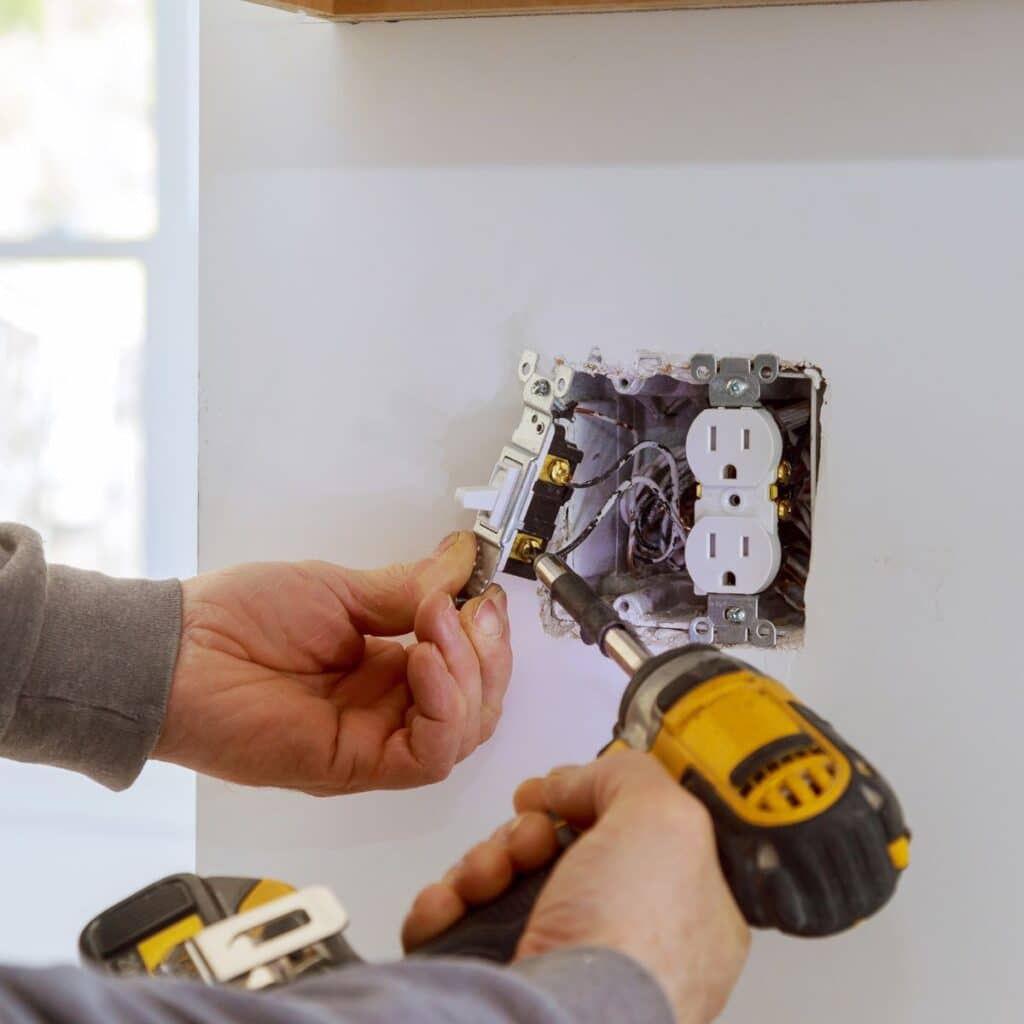 A person's hands are shown wiring an electrical socket, with one hand holding a screwdriver and another preparing the wiring. The focus on the hands against the backdrop of the open electrical box illustrates precise and skilled work, likely during a home renovation or electrical maintenance task