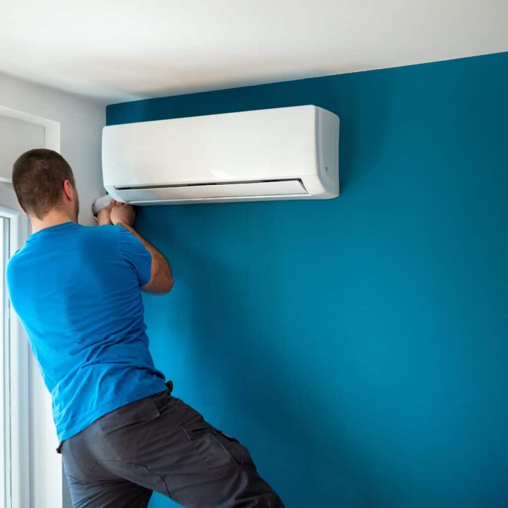 A person in a blue shirt is installing or repairing a white air conditioning unit on a vibrant blue wall. The individual's focus is on the task at hand, as they reach up to adjust or fix something on the bottom of the unit
