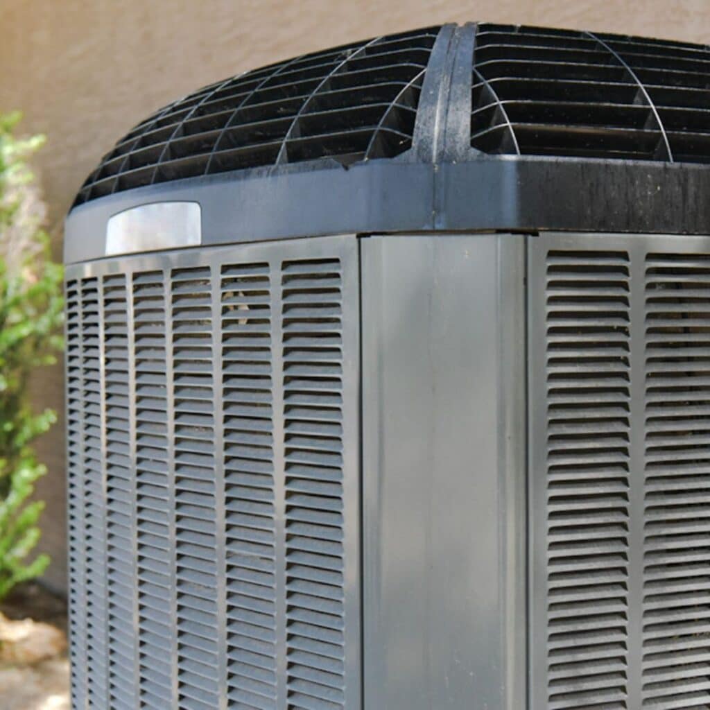 A close-up of a residential central air conditioning unit outside a home. The focus on the fan grid and the unit's casing highlights the importance of this equipment in home climate control