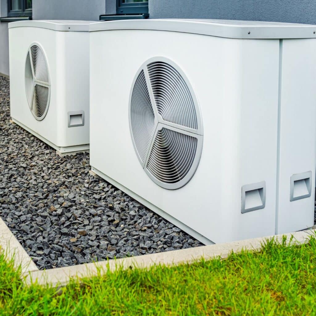 Two large, white external HVAC units with prominent circular fans are positioned on a bed of gray gravel next to a building, bordered by a well-maintained grassy area. The design suggests a modern and efficient air circulation system for climate control within the premises