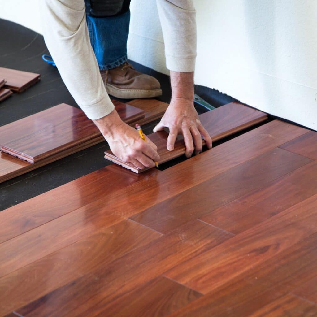 A person is kneeling on the floor, installing dark wood laminate flooring in a room. The individual's hands are visible, marking a piece of the laminate with a pencil to ensure precise fitting. This task indicates a home improvement project focusing on flooring installation