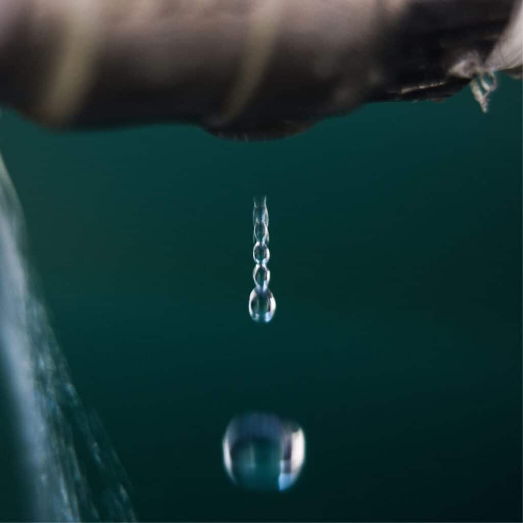 A macro shot capturing the moment water drips from a faucet, with each droplet in sharp focus against a blurred green background. The image conveys the theme of water conservation or plumbing issues, emphasizing the clarity and preciousness of water