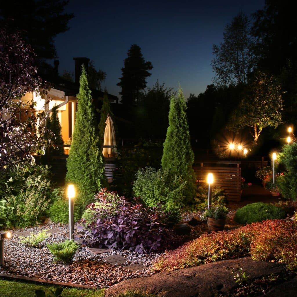 An enchanting nighttime garden illuminated by soft, warm lighting. Tall, narrow conifer trees rise above a well-manicured landscape, with various shrubs and plants creating a textured tapestry of greens and purples. The garden's design features a meandering path lined with decorative stones and punctuated by round, glowing lights that cast a tranquil ambiance. In the background, the edge of a cozy home peeks through the foliage, suggesting a serene domestic outdoor setting