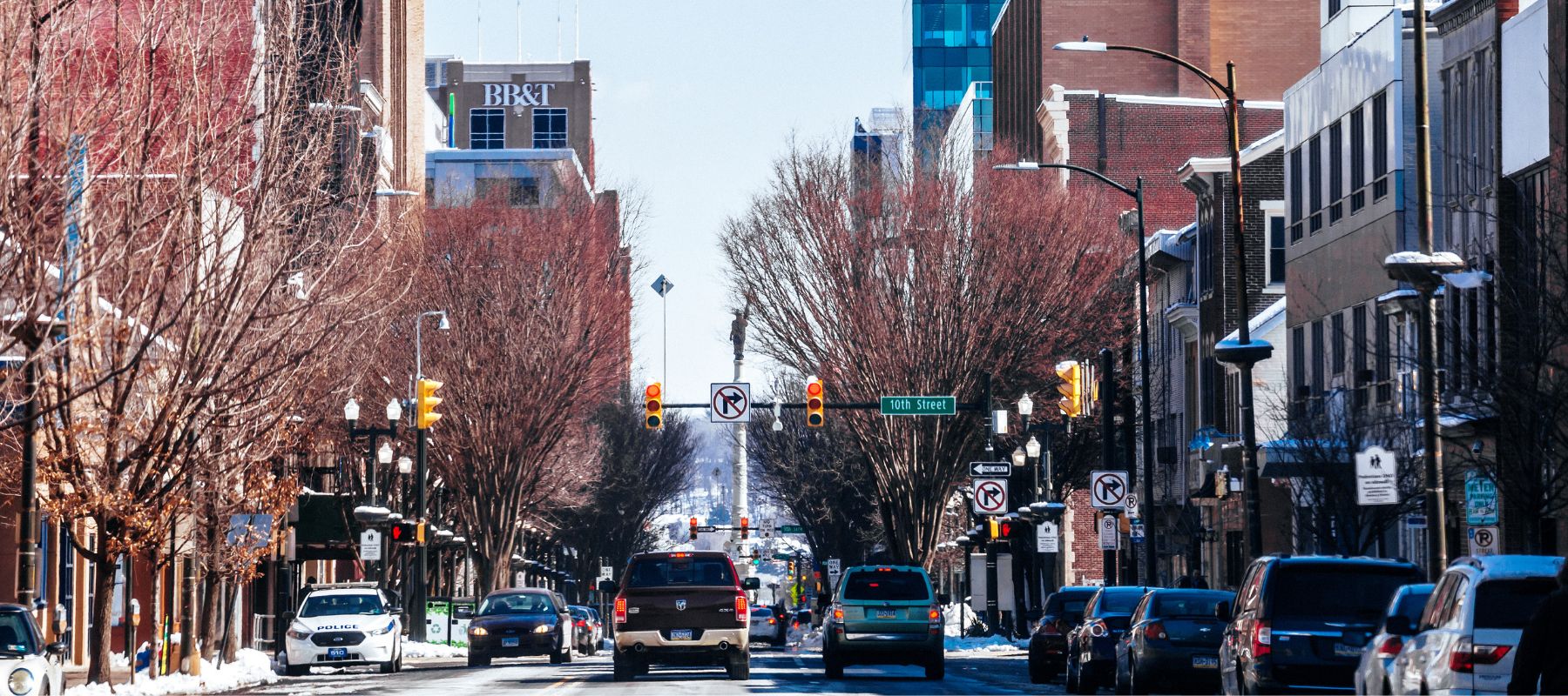 A bustling city street lined with leafless trees and buildings, including a bank, under a clear sky. The image captures the urban rhythm with cars, street lights, and pedestrians, framing a typical day in a downtown area during winter