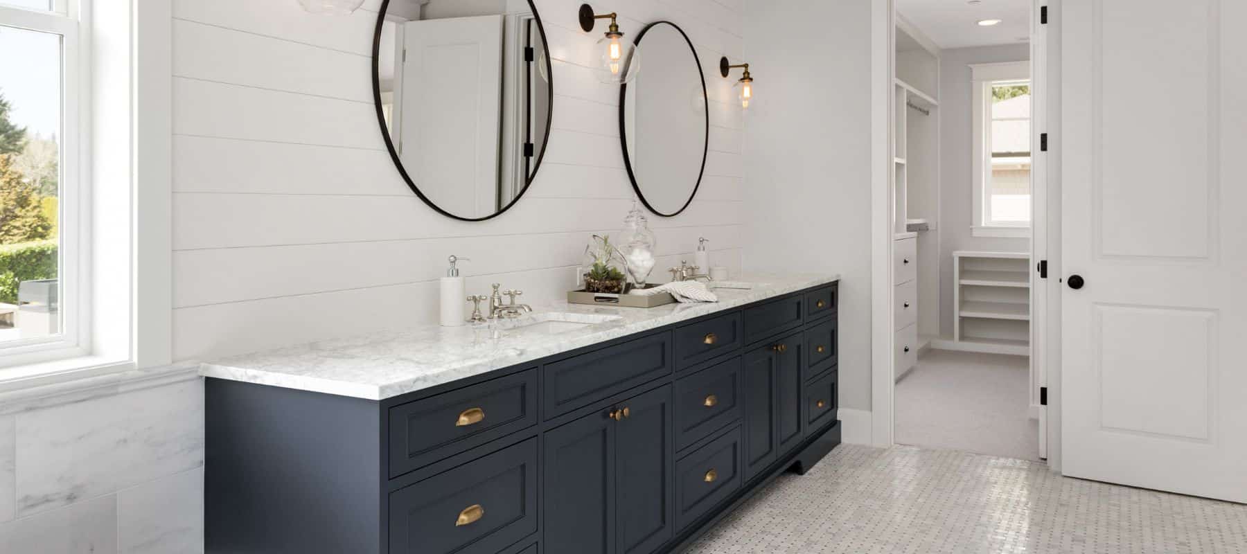 A stylish bathroom interior featuring a long vanity with dark navy cabinets and a white marble countertop. Above the vanity, two large round mirrors with black frames are hung on a white shiplap wall, flanked by elegant wall-mounted light fixtures with exposed bulbs. The floor is tiled with small white hexagonal pieces, and a glimpse of a built-in white closet adds to the clean and modern aesthetic of the space