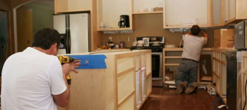 Two individuals are focused on a kitchen remodeling project. One person is drilling into a section of cabinetry covered with blue tape, while another is working in the background, demonstrating the collaborative effort involved in home improvement