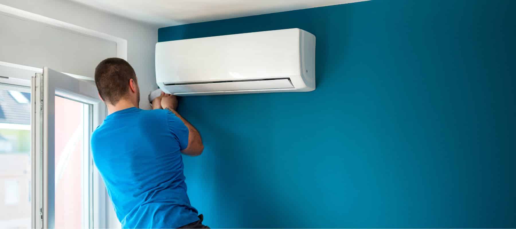 A person in a blue shirt is installing or repairing a white air conditioning unit on a vibrant blue wall. The individual's focus is on the task at hand, as they reach up to adjust or fix something on the bottom of the unit