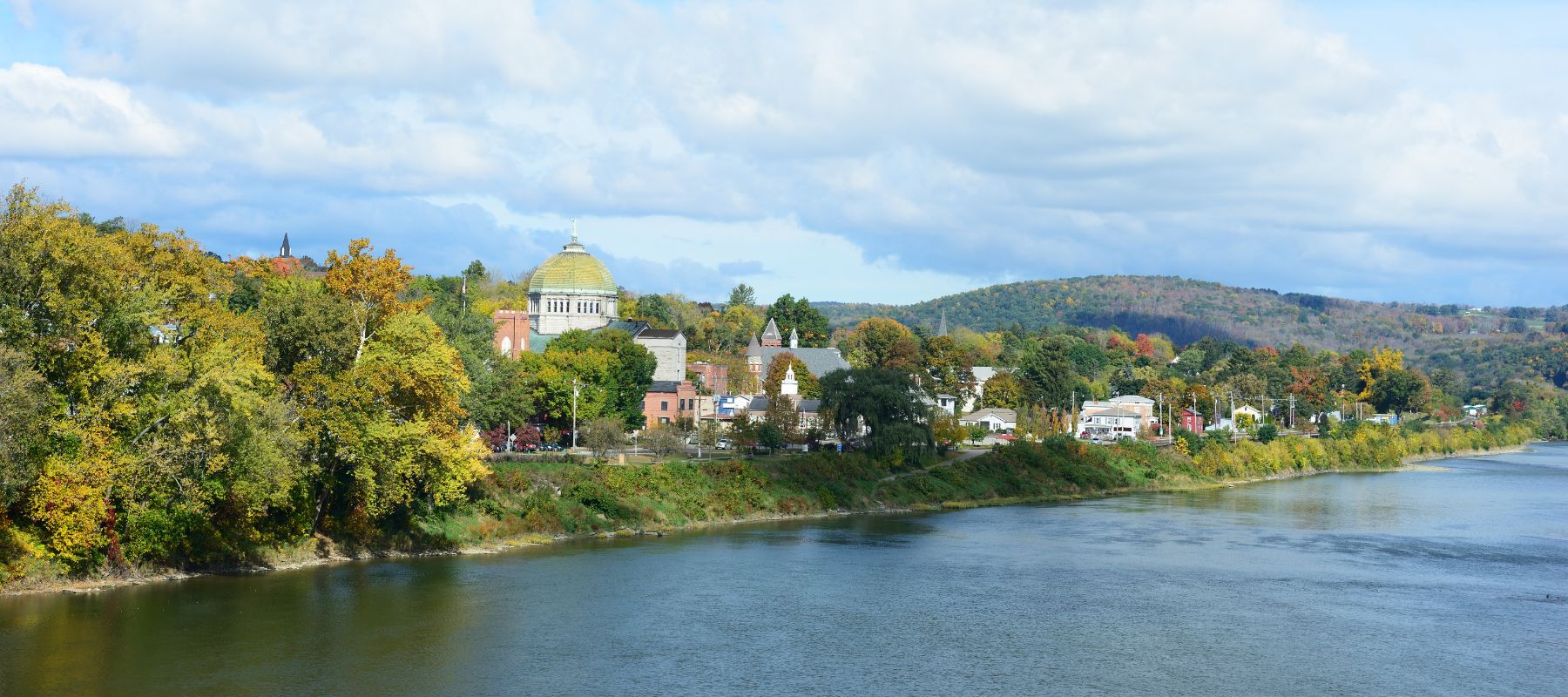 A sweeping view of a picturesque riverside town during fall, with colorful foliage and a grand domed building as the centerpiece. The scene reflects a tranquil, small-town atmosphere with natural beauty and historic architecture