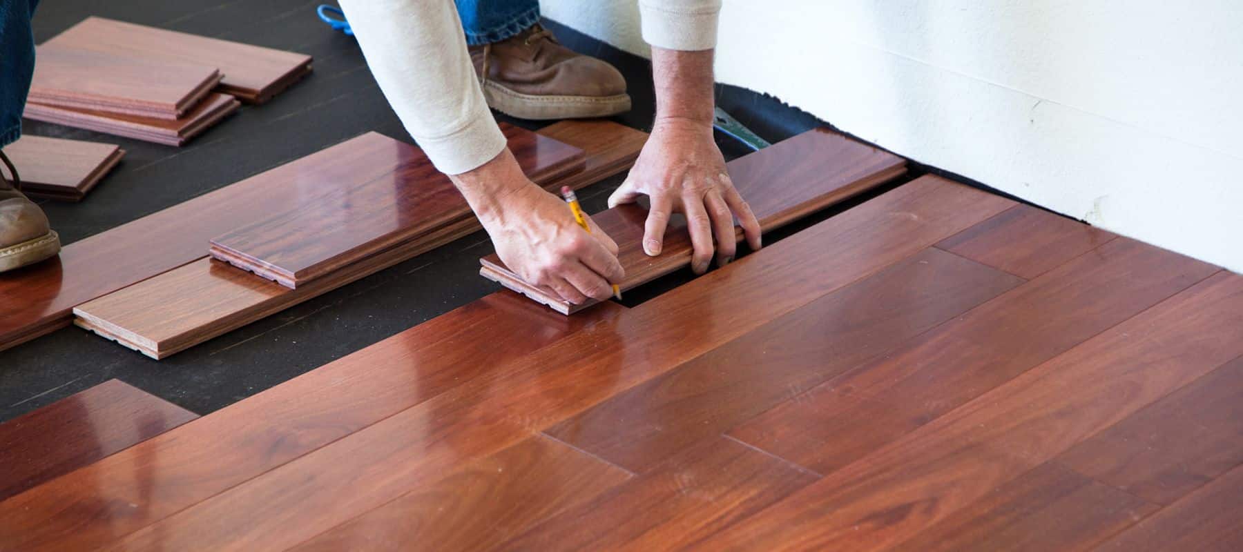 A person is kneeling on the floor, installing dark wood laminate flooring in a room. The individual's hands are visible, marking a piece of the laminate with a pencil to ensure precise fitting. This task indicates a home improvement project focusing on flooring installation