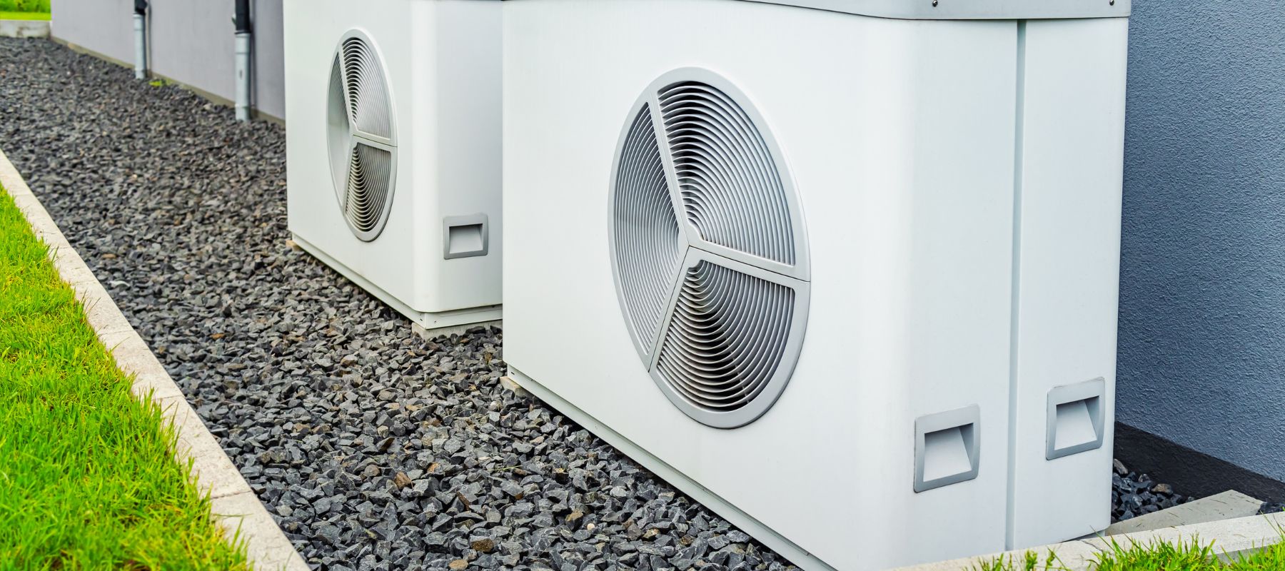Two large, white external HVAC units with prominent circular fans are positioned on a bed of gray gravel next to a building, bordered by a well-maintained grassy area. The design suggests a modern and efficient air circulation system for climate control within the premises