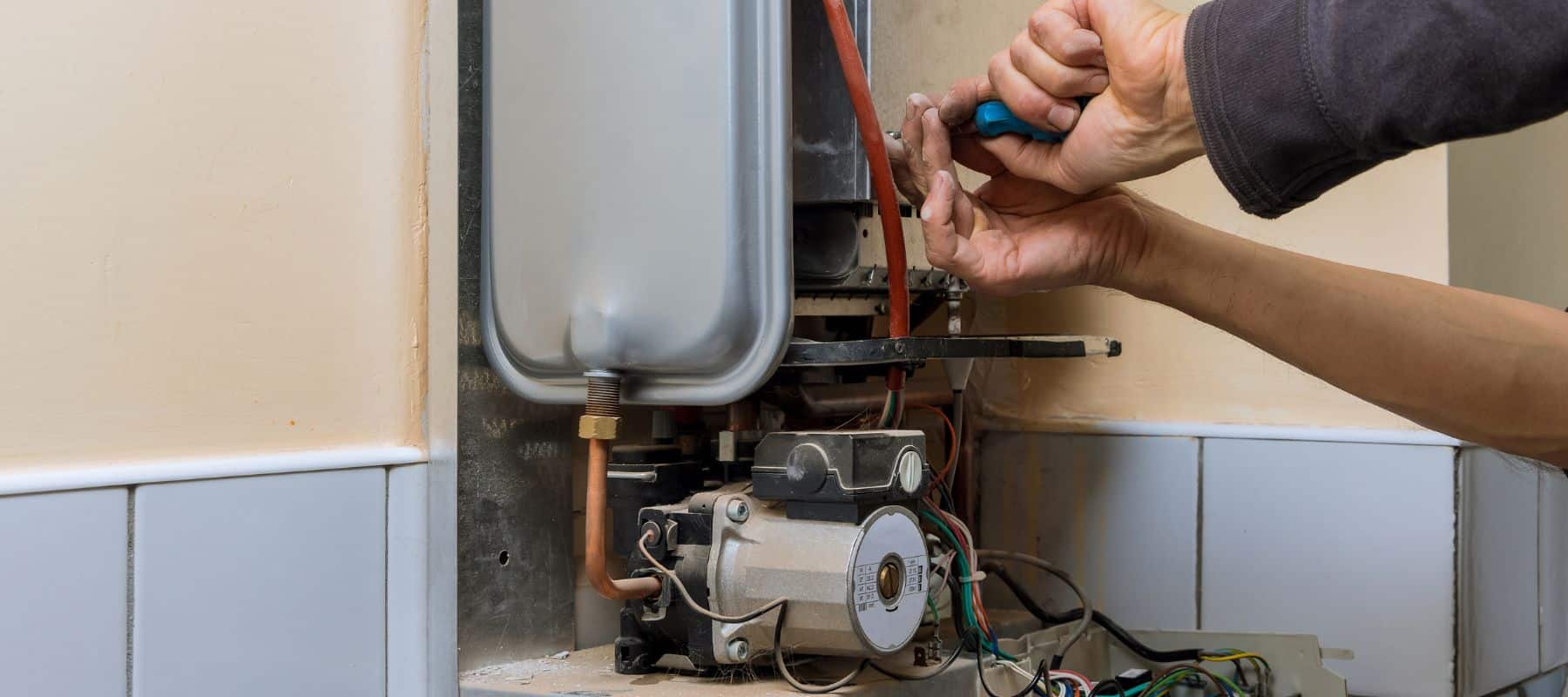 A person is servicing a boiler or water heater, visible by their hands using a screwdriver to adjust components within the lower service panel. The complexity of the machinery, with various wires and pipes, suggests technical expertise is required for the task. The maintenance work is being performed in a domestic setting, indicated by the tiled wall and flooring
