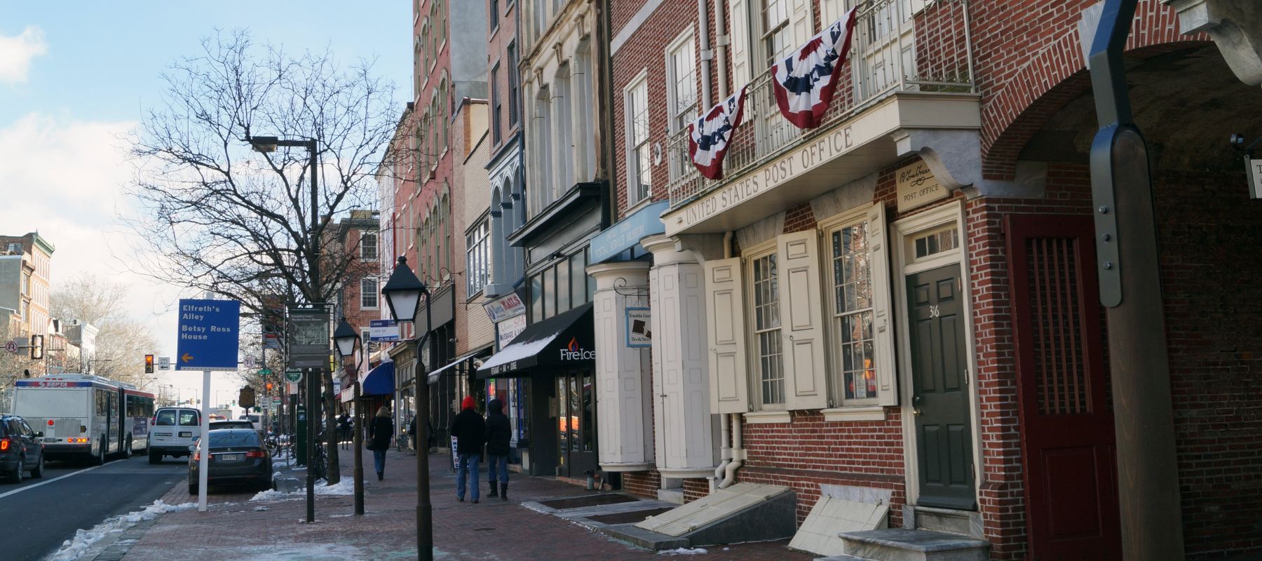 A panoramic view of a historic city street with traditional brick buildings, American flags, and a clear winter sky. Pedestrians and vehicles suggest everyday urban activity, while the signage points towards landmarks like the Betsy Ross House, infusing the scene with a sense of heritage and continuity in north wales pennsylvania