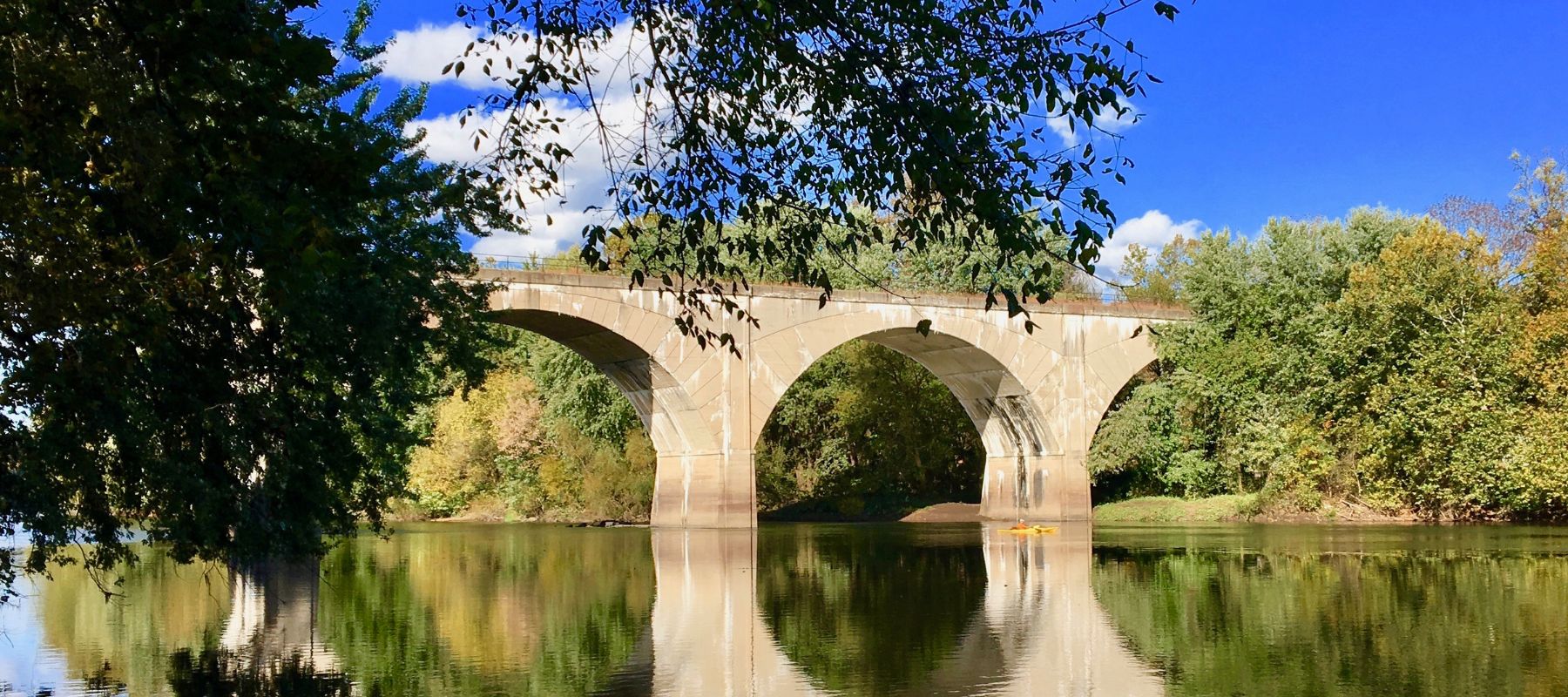 A tranquil scene of an arched stone bridge over a calm river, reflecting the bridge's silhouette. The lush greenery on the banks frames the view, and a clear blue sky above completes this picturesque setting
