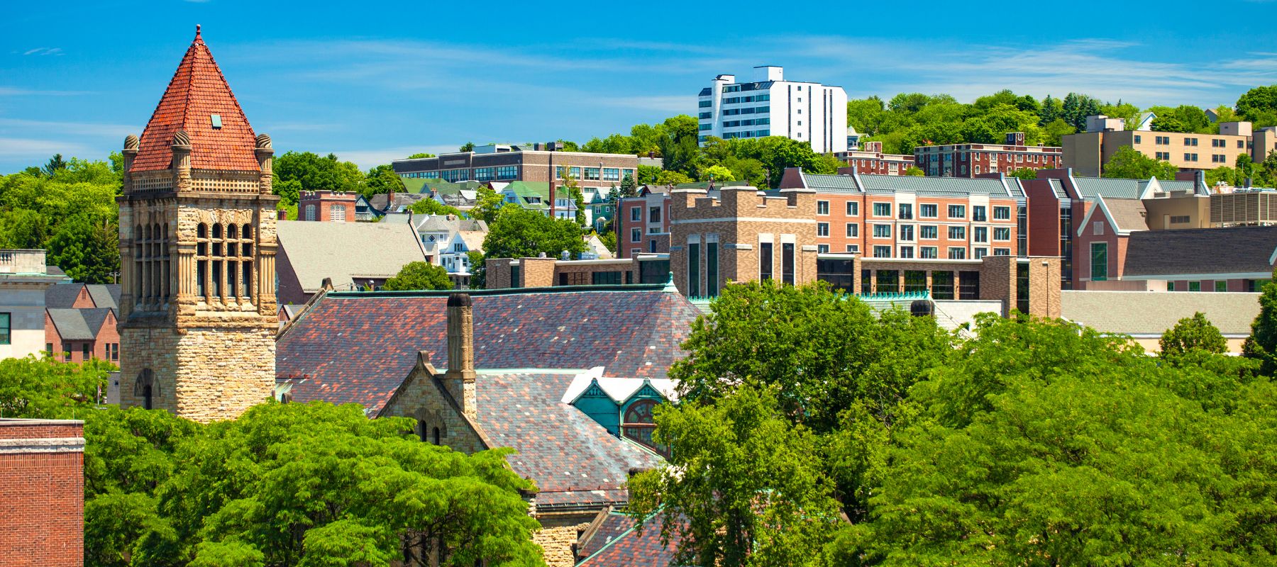 A vibrant cityscape with diverse architectural styles, including a prominent stone tower with a conical roof, modern buildings, and classic homes nestled among lush green trees under a bright blue sky
