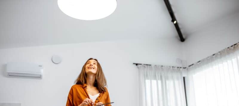 A happy woman in a terracotta-colored shirt is indoors, looking up towards a large circular skylight that floods the room with natural light. To her side, a modern air conditioning unit is mounted on the wall, indicating a comfortable and well-ventilated space