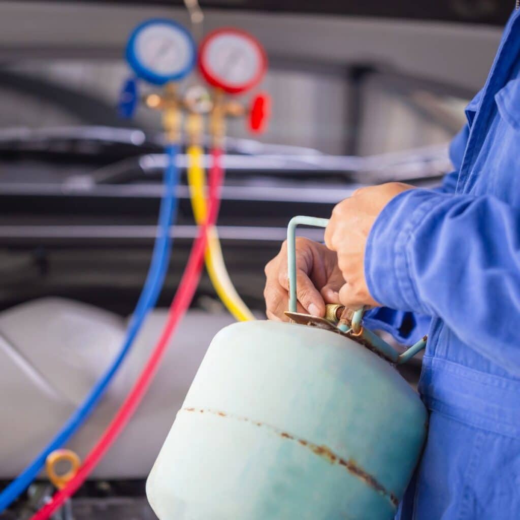 A technician in blue workwear is handling a refrigerant tank with gauges and hoses attached, likely performing maintenance or charging an air conditioning system. The focus on the hands and tools foregrounds the precision required in HVAC services