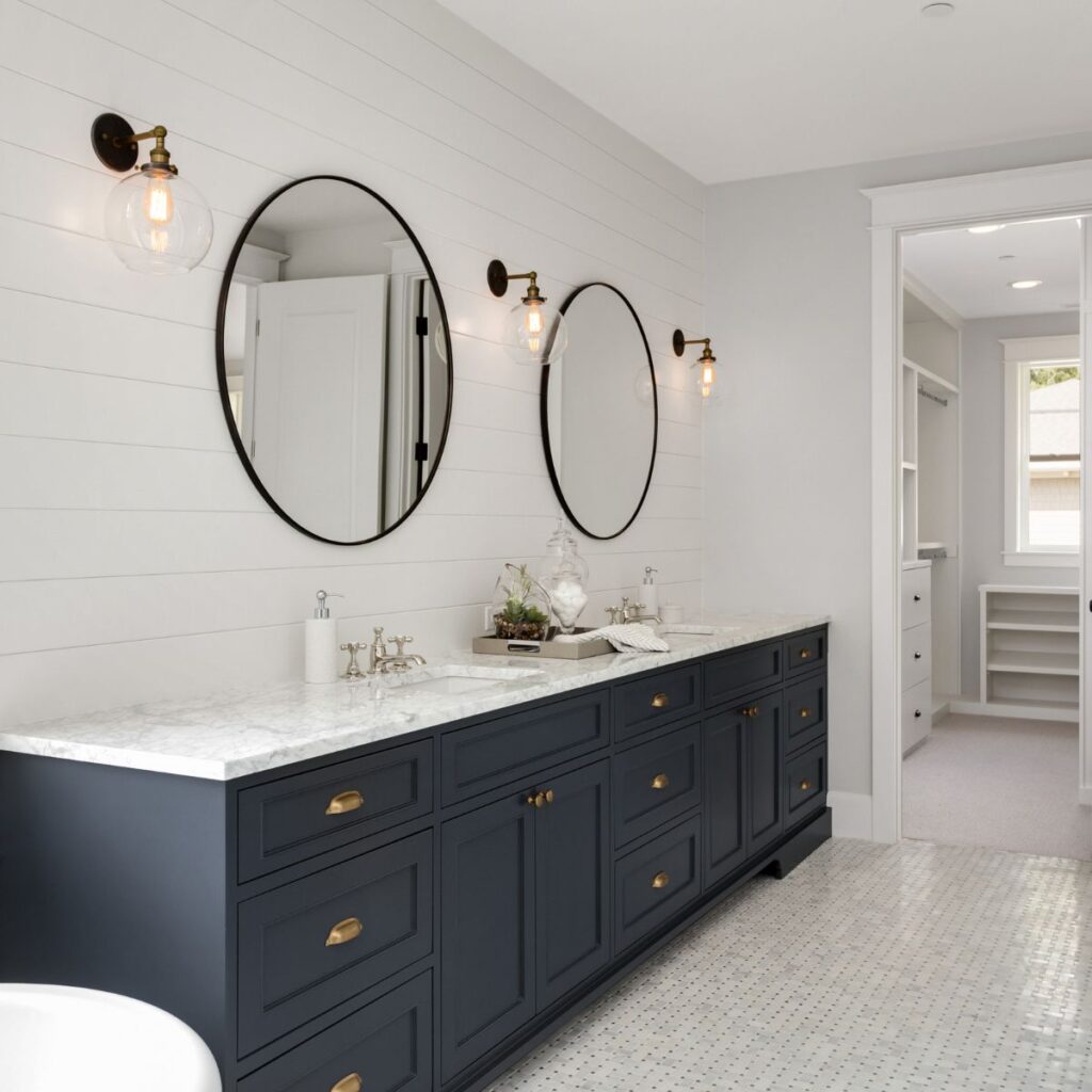 A stylish bathroom interior featuring a long vanity with dark navy cabinets and a white marble countertop. Above the vanity, two large round mirrors with black frames are hung on a white shiplap wall, flanked by elegant wall-mounted light fixtures with exposed bulbs. The floor is tiled with small white hexagonal pieces, and a glimpse of a built-in white closet adds to the clean and modern aesthetic of the space