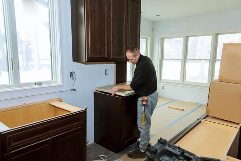 A man in a dark long-sleeve shirt and jeans is installing a dark brown wooden kitchen cabinet in a room with natural light. The room has several cabinets and countertop pieces, and blue painter's tape on the floor, indicating ongoing remodeling or installation work