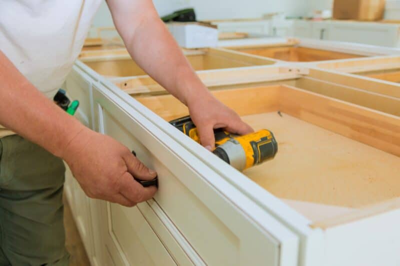 Close-up view of a person installing a drawer using a cordless drill into a white, wooden kitchen cabinet frame. The scene focuses on the worker's hands as they carefully secure the drawer, highlighting the detail and precision involved in cabinet installation