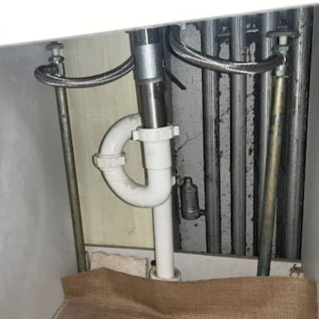 Under-sink plumbing with a white P-trap pipe and metal supply lines against a wall with multiple vertical pipes