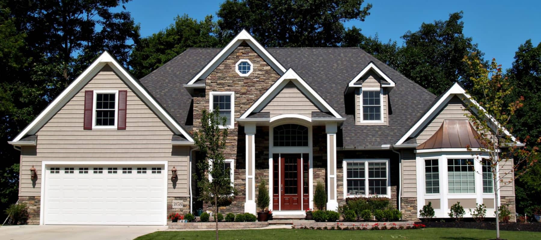 Elegant home with a mix of tan siding and stone detailing, white trim, and a manicured front lawn