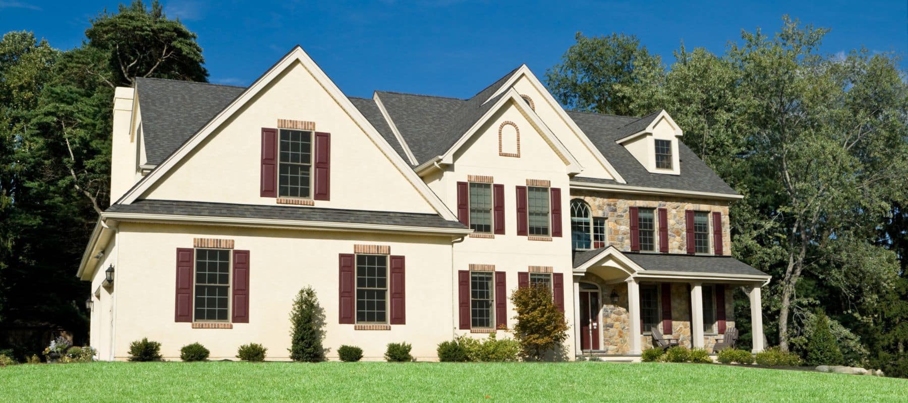 Large house with cream-colored stucco, maroon shutters, and partial stone facade surrounded by tall trees and green lawn