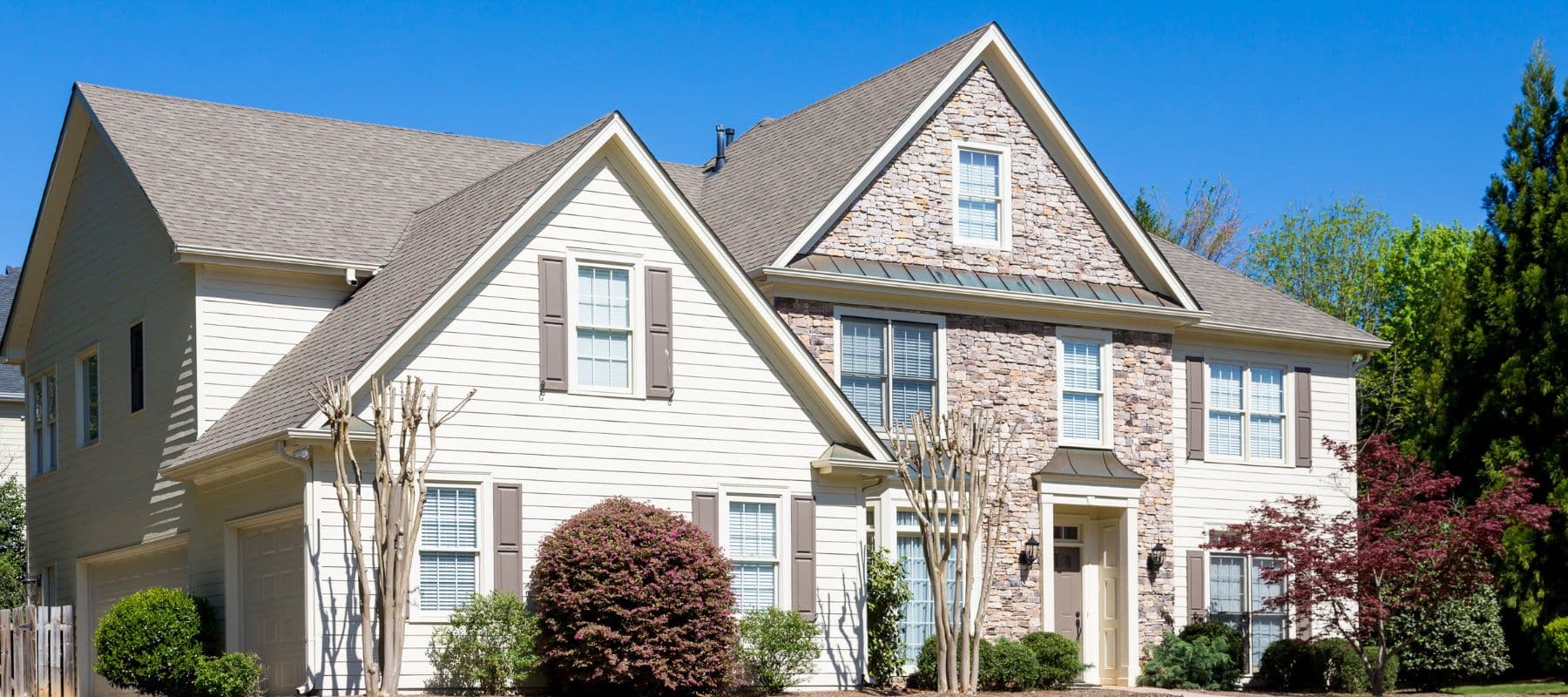 Spacious light beige home with taupe shutters and a prominent stone facade, surrounded by lush greenery