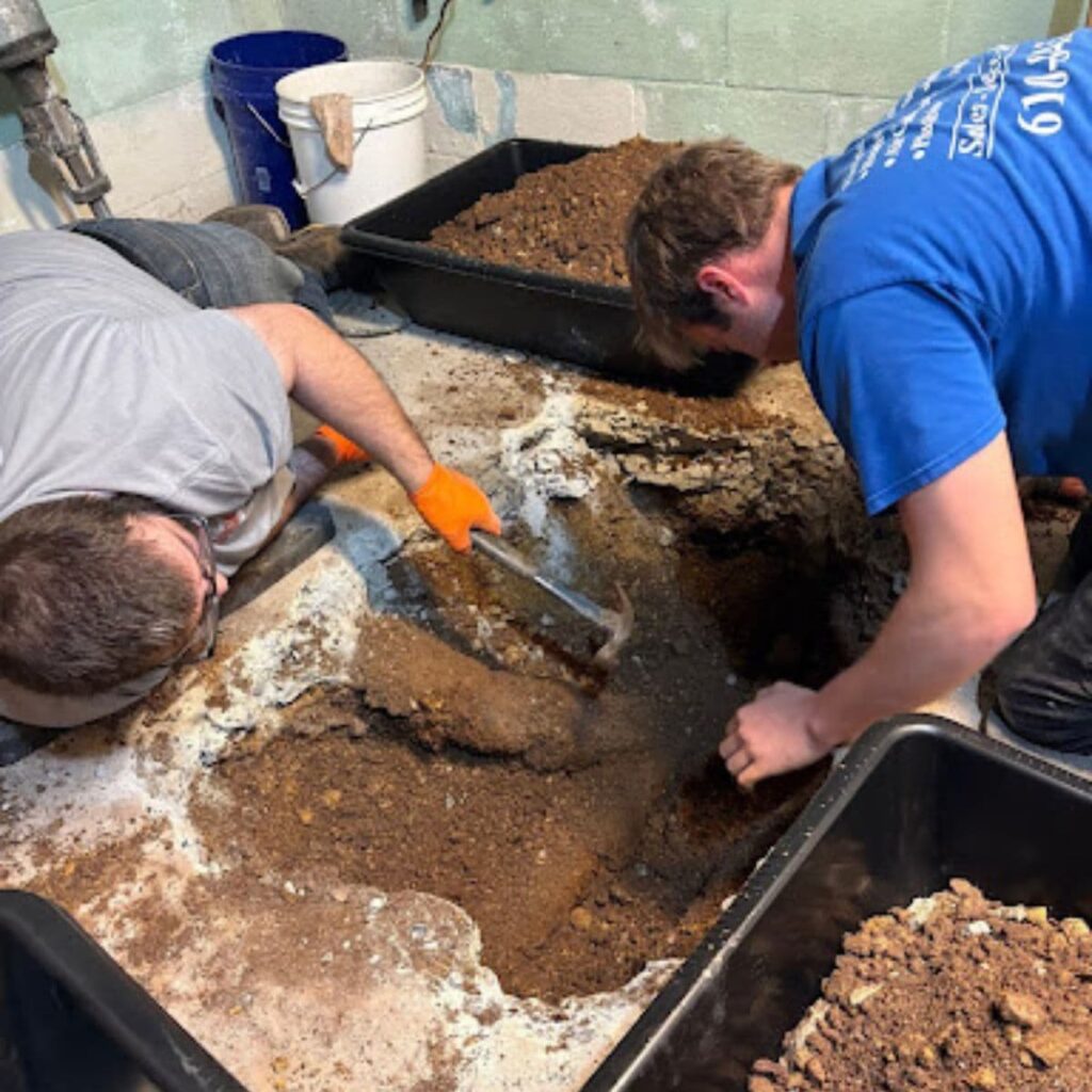Two plumbers dig through a concrete floor and dirt in a basement to access underground plumbing