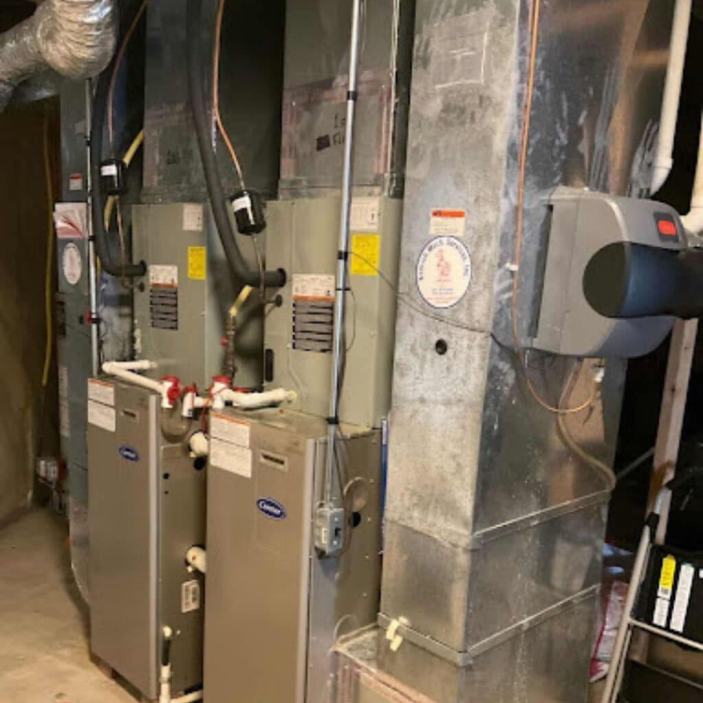 Two indoor furnace systems with visible piping and ductwork installed side-by-side in a mechanical room