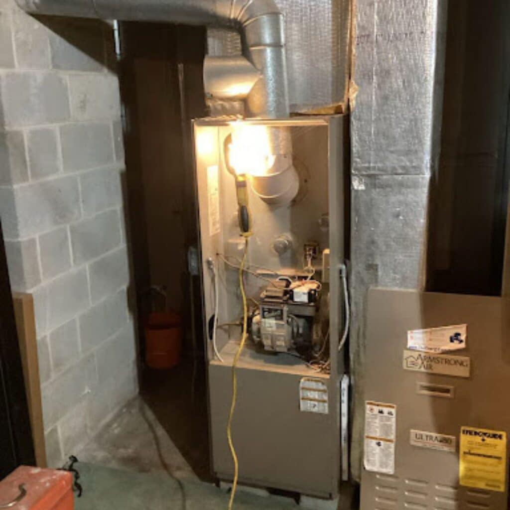 An open furnace unit being serviced in a basement, with tools and wiring exposed under a bright work light
