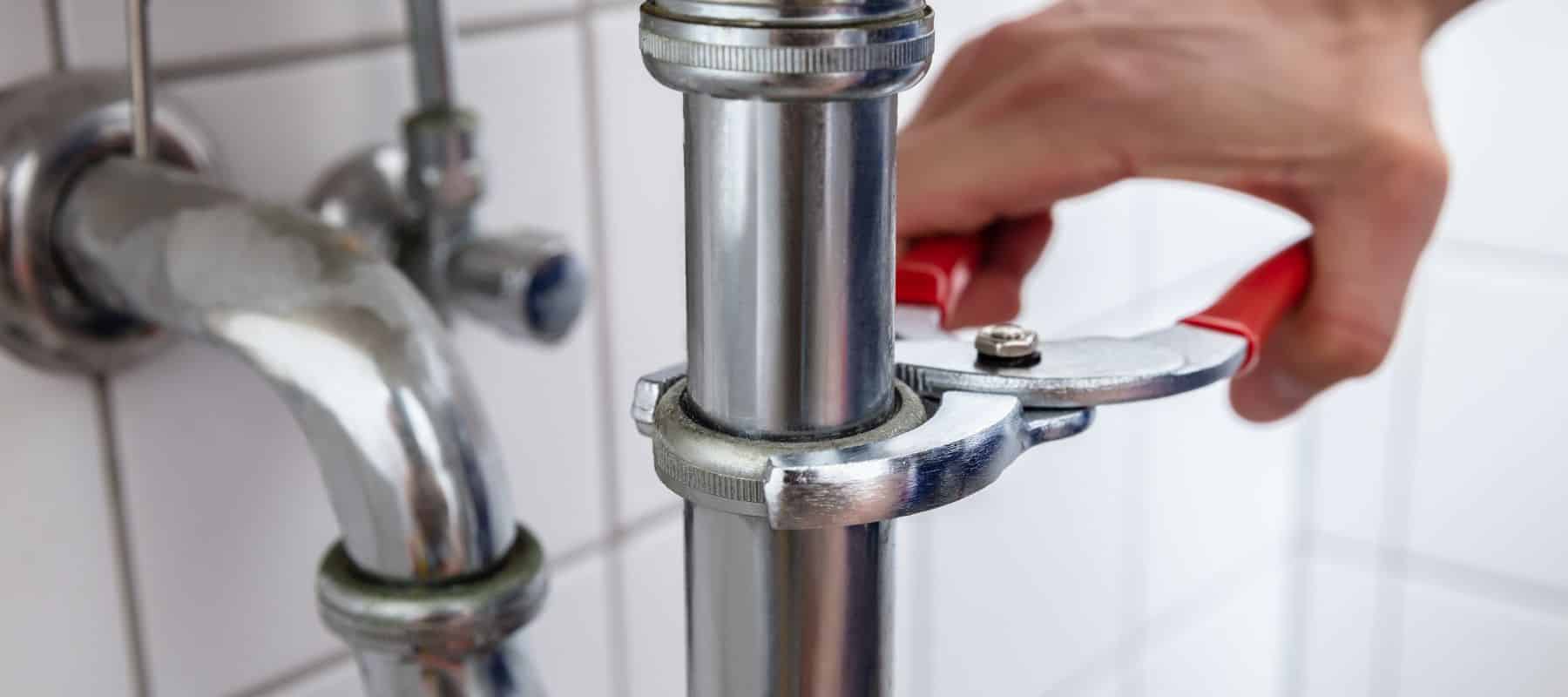 A hand uses a red-handled wrench to tighten a connection on a chrome pipe under a sink