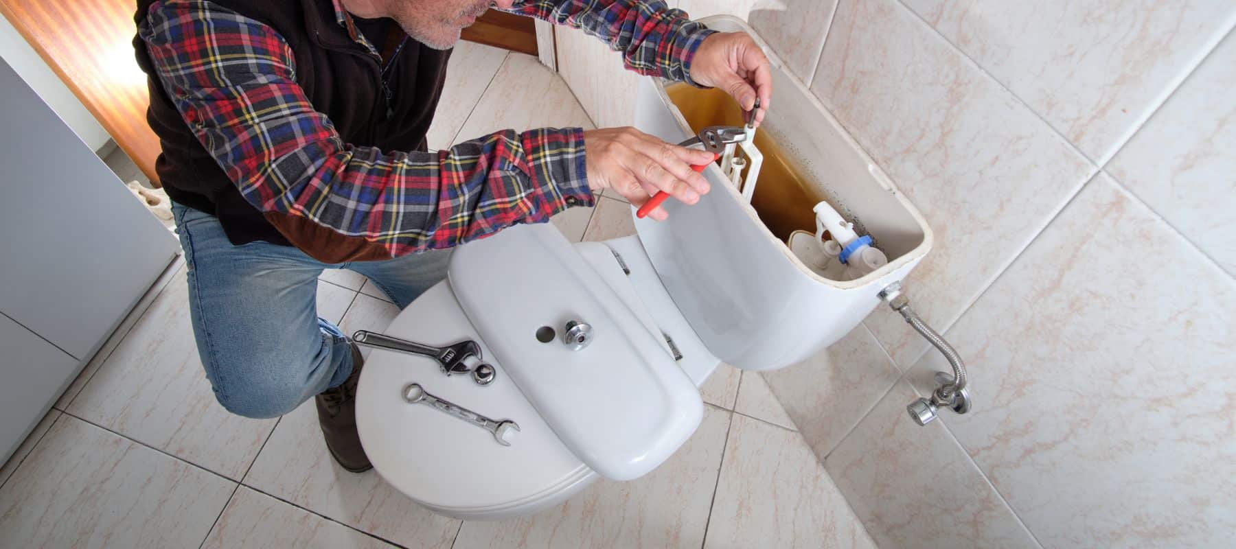 A plumber uses a wrench to work on the internal mechanisms of an open toilet tank from a high angle