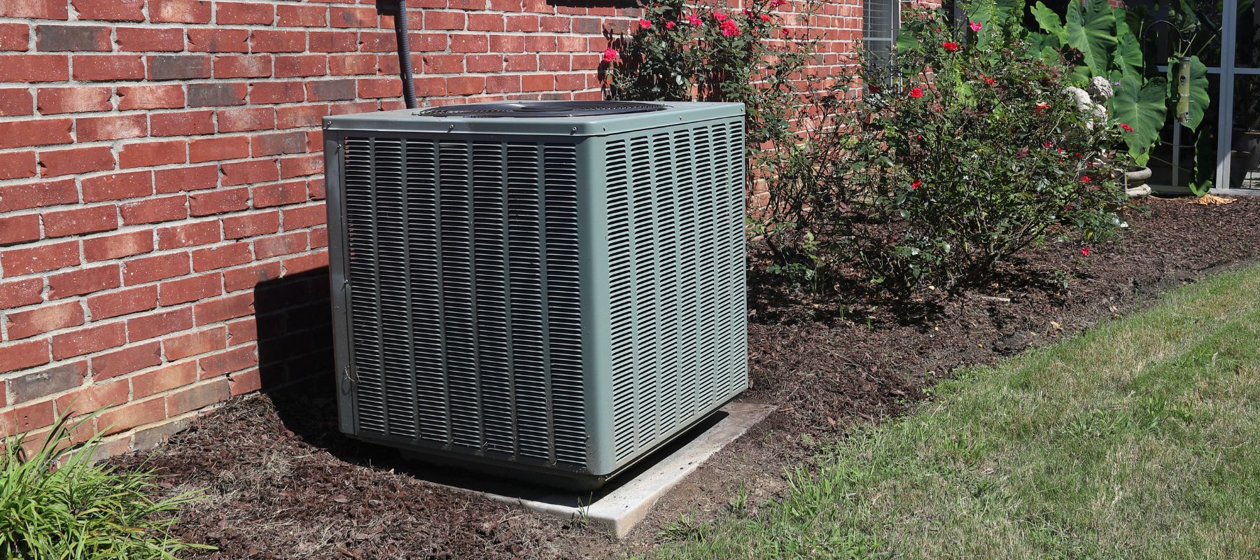 A large outdoor air conditioning unit sits on a concrete pad next to a brick house, surrounded by green grass and landscaping