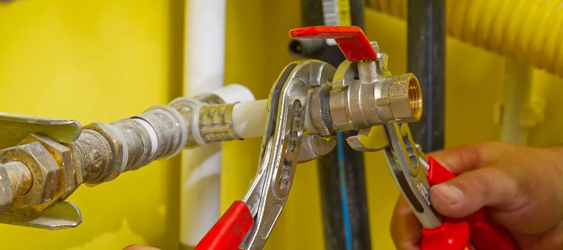 A close-up shows hands using two wrenches to work on a metal pipe with a red valve, set against a yellow background