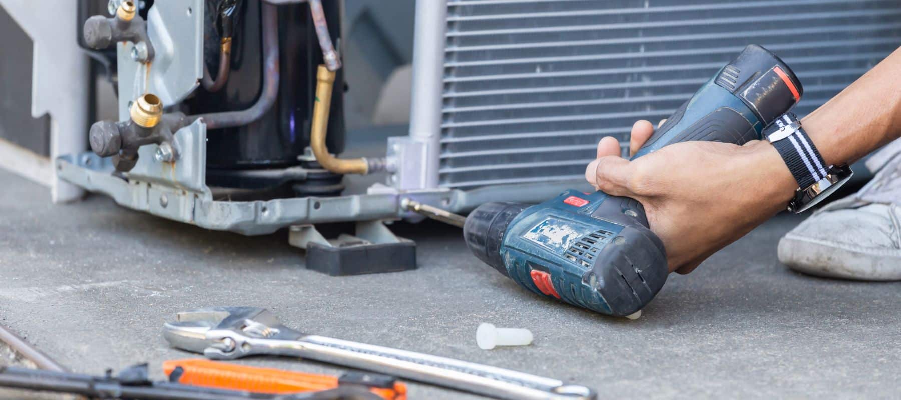 A person's hands use a power drill to work on the internal components of an outdoor HVAC unit