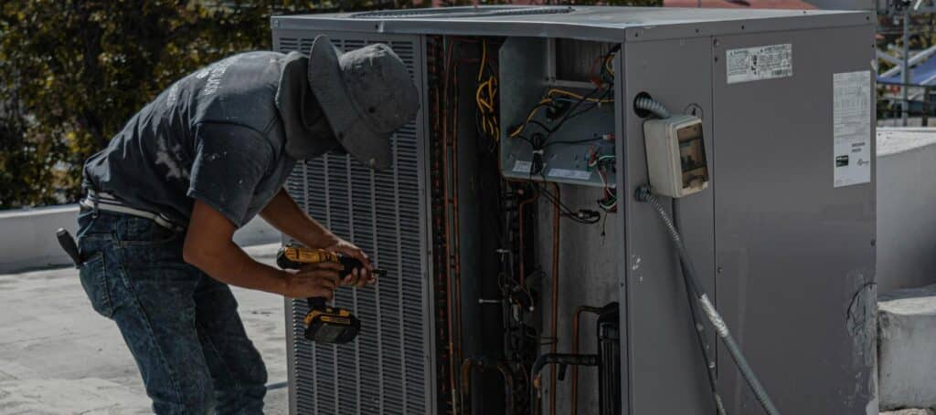 An HVAC technician in a hat and work clothes uses a power drill to service an outdoor air conditioning unit