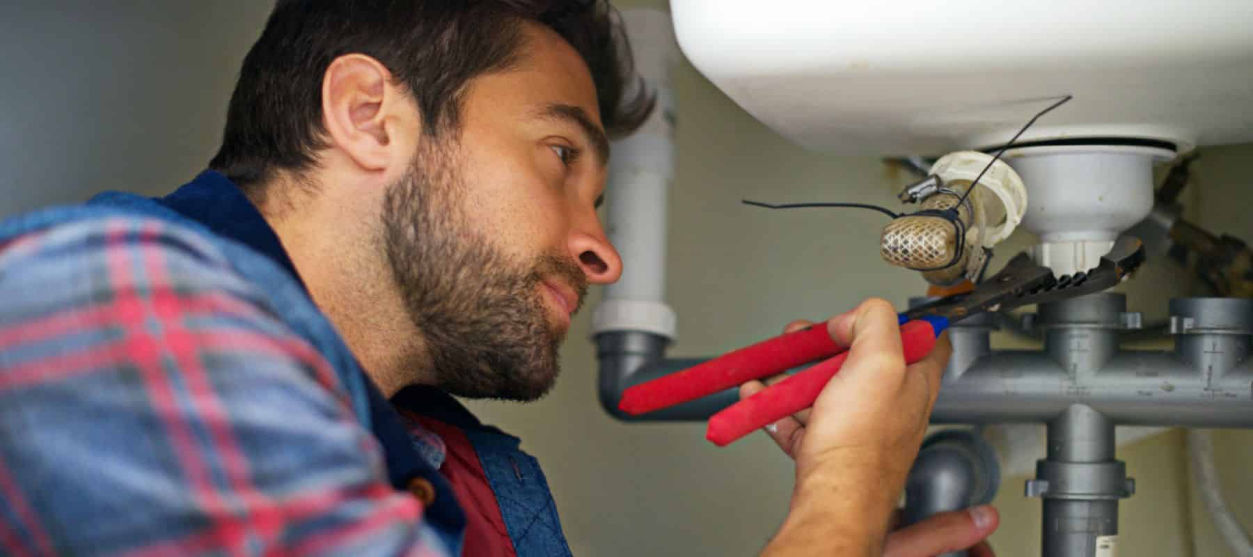 A bearded plumber, looking intently, uses pliers to work on the pipes beneath a sink