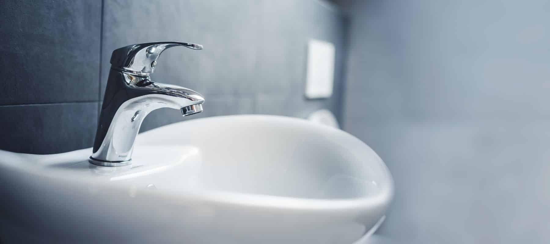 A gleaming chrome faucet is prominently featured over a white sink basin, with a blurred dark tiled wall in the background