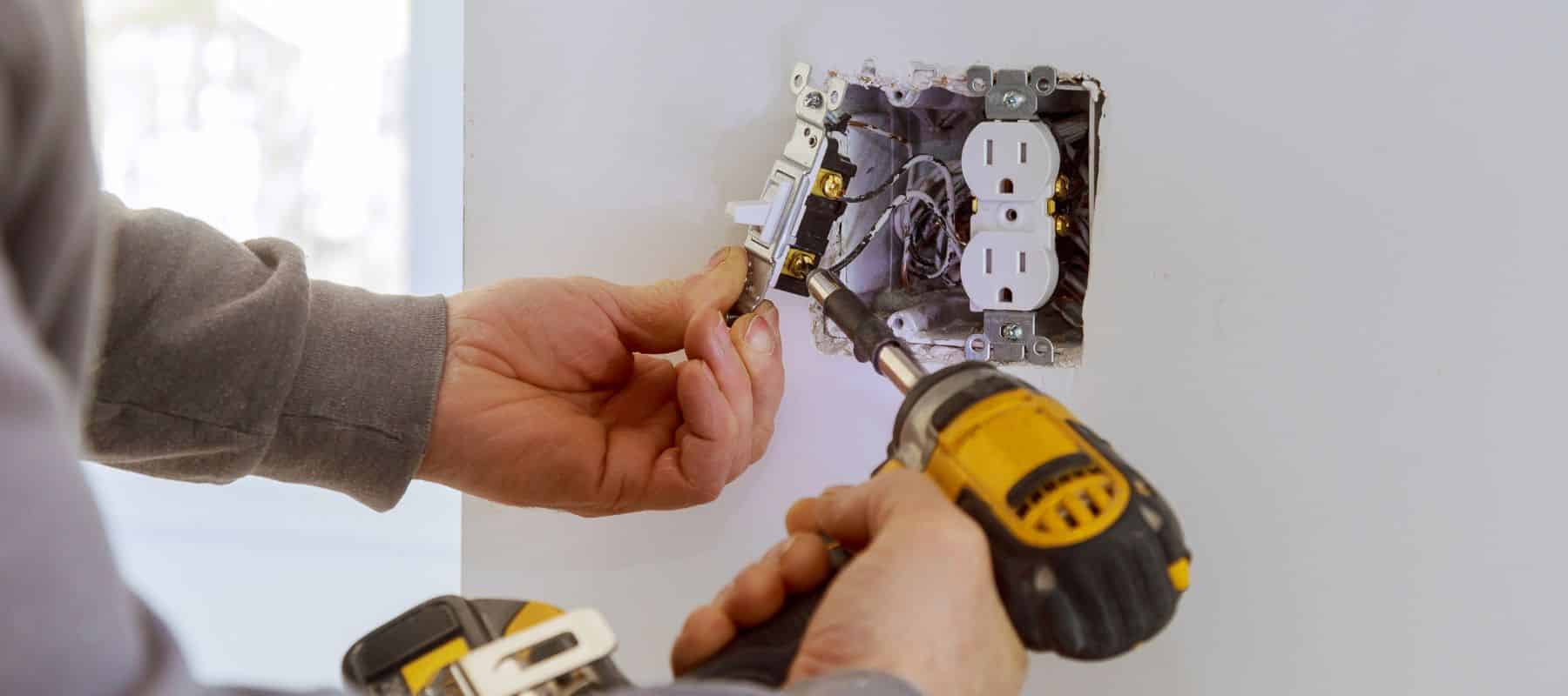 An electrician uses a power drill to install a white electrical outlet and switch in a wall